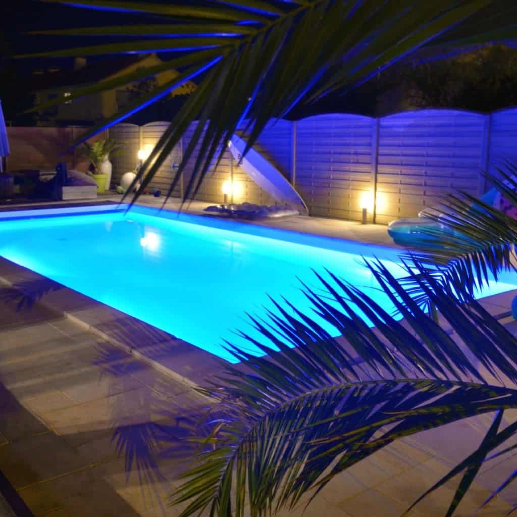 A swimming pool illuminated with bright blue lights during nighttime, surrounded by palm leaves and a privacy fence. Soft yellow lights enhance the serene atmosphere, creating a relaxing outdoor setting.