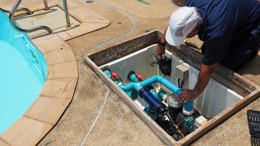 A technician in a navy shirt and white cap is inspecting and repairing pool pump equipment in an open maintenance box beside a swimming pool. The equipment includes blue pipes and various components.