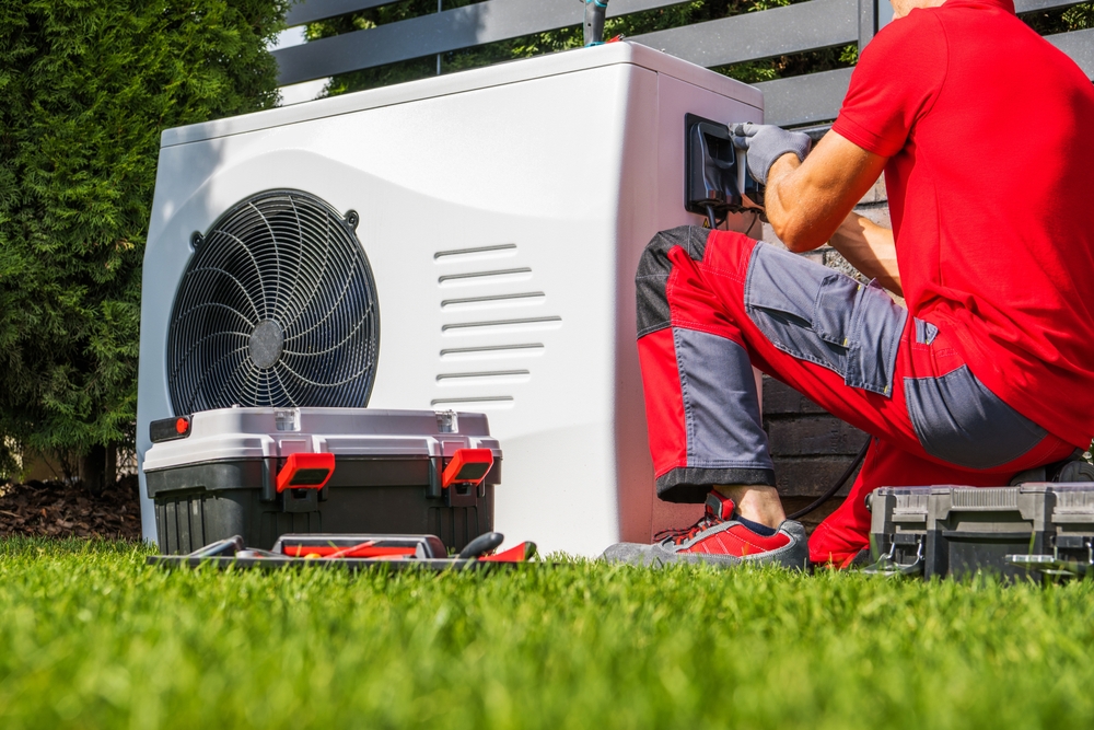 A technician in red workwear repairs or installs a large outdoor generator or HVAC unit in a grassy yard, with toolboxes placed nearby.