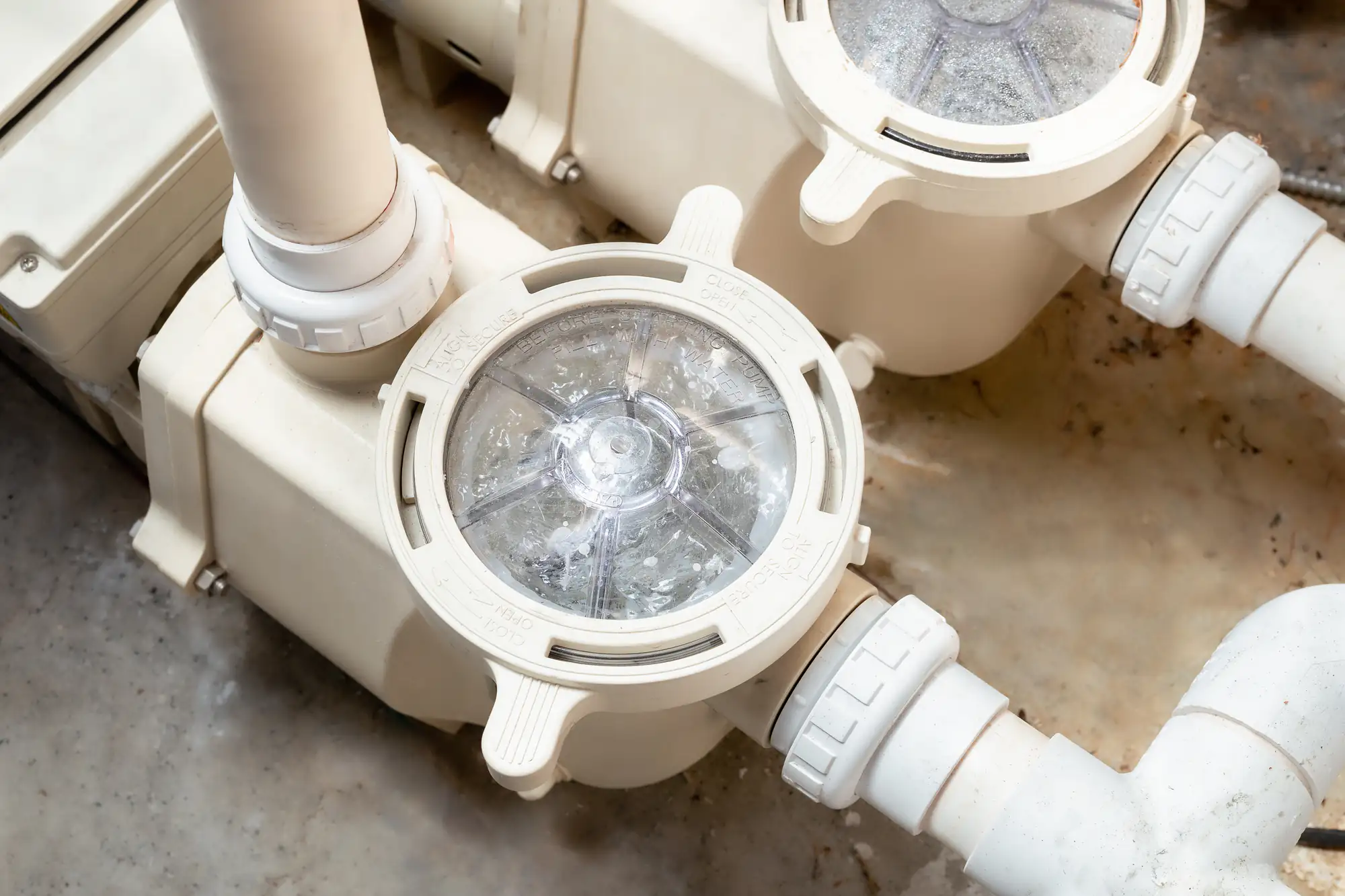Close-up of a swimming pool pump with a transparent lid, showing water swirling inside. White pipes are connected to the pump, which is mounted on a concrete surface.