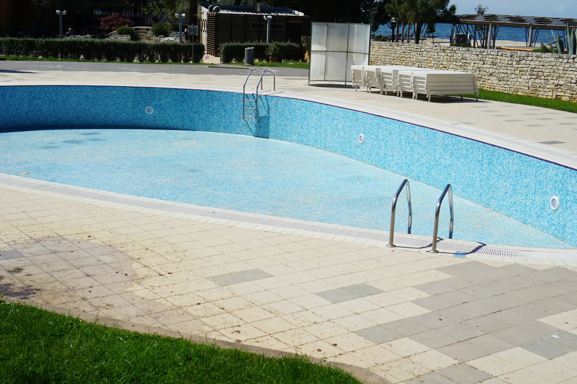 An empty outdoor swimming pool with blue tiles, surrounded by a tiled deck. Metal ladders are on each end, and white lounge chairs are arranged nearby. Grass borders part of the pool deck.