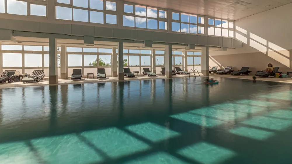 Indoor swimming pool with clear water, surrounded by lounge chairs and large windows letting in abundant sunlight. A few people are relaxing on the chairs, and one person is floating in the pool.