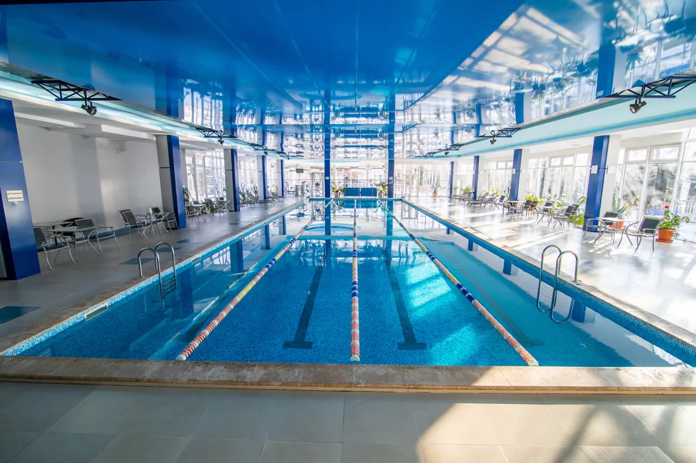Indoor swimming pool with clear blue water, lane dividers, and ladders on both sides. The room has a shiny blue ceiling, large windows, and seating areas with tables and chairs along the sides.
