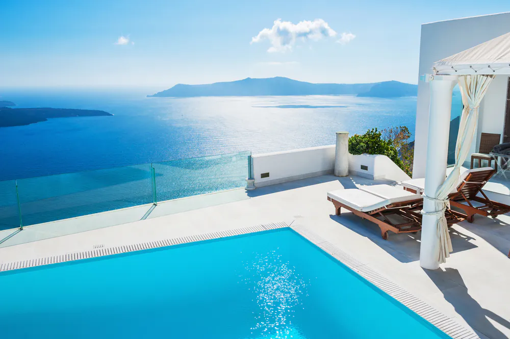 A bright infinity pool overlooks a calm blue sea with distant islands under a clear sky; two wooden lounge chairs and a pergola sit on a white patio, creating a relaxing, luxurious atmosphere.