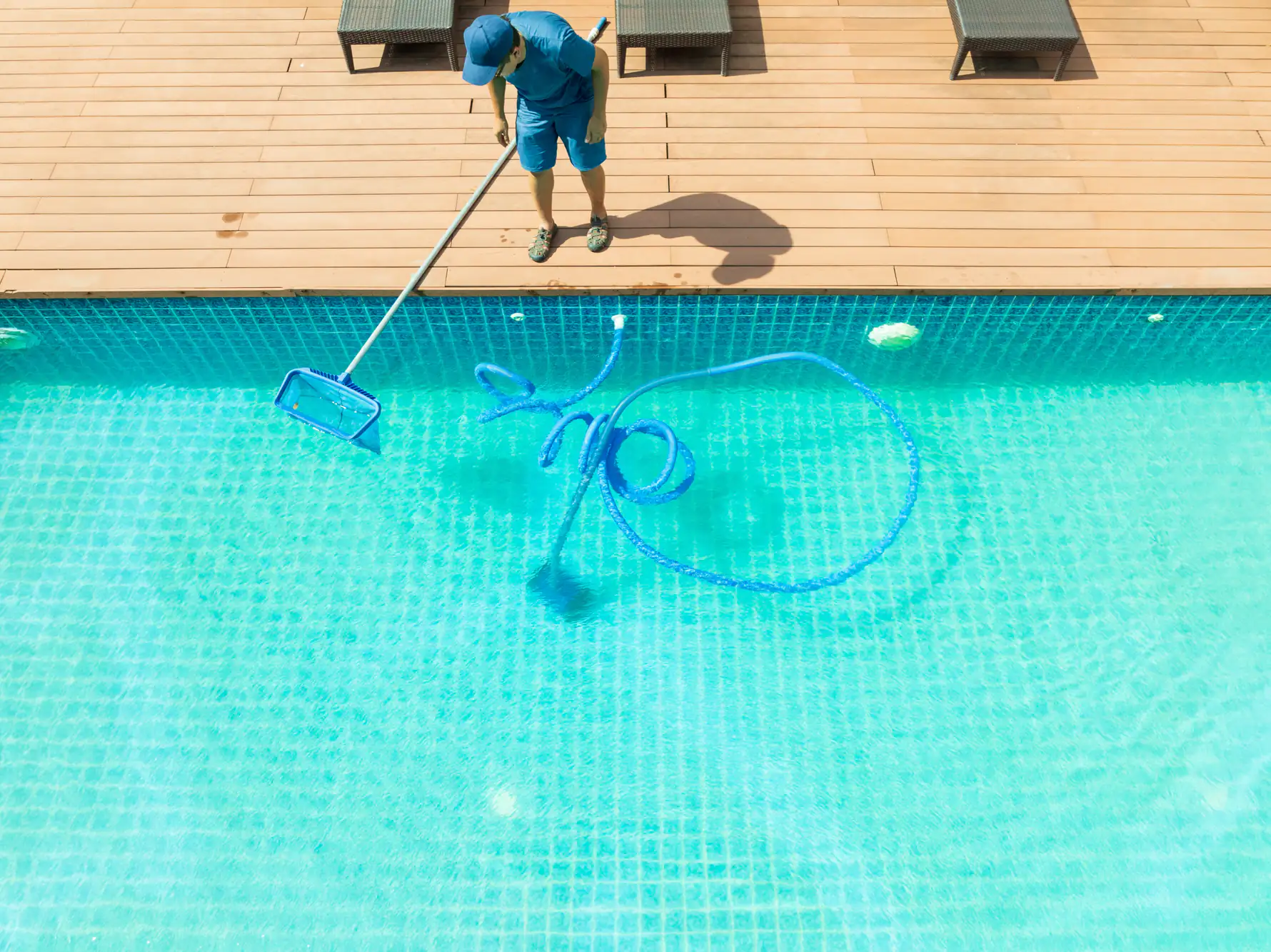 A person in blue clothes and cap is cleaning a clear swimming pool with a net on a pole, standing on a wooden deck beside lounge chairs, with a blue hose coiled in the water.