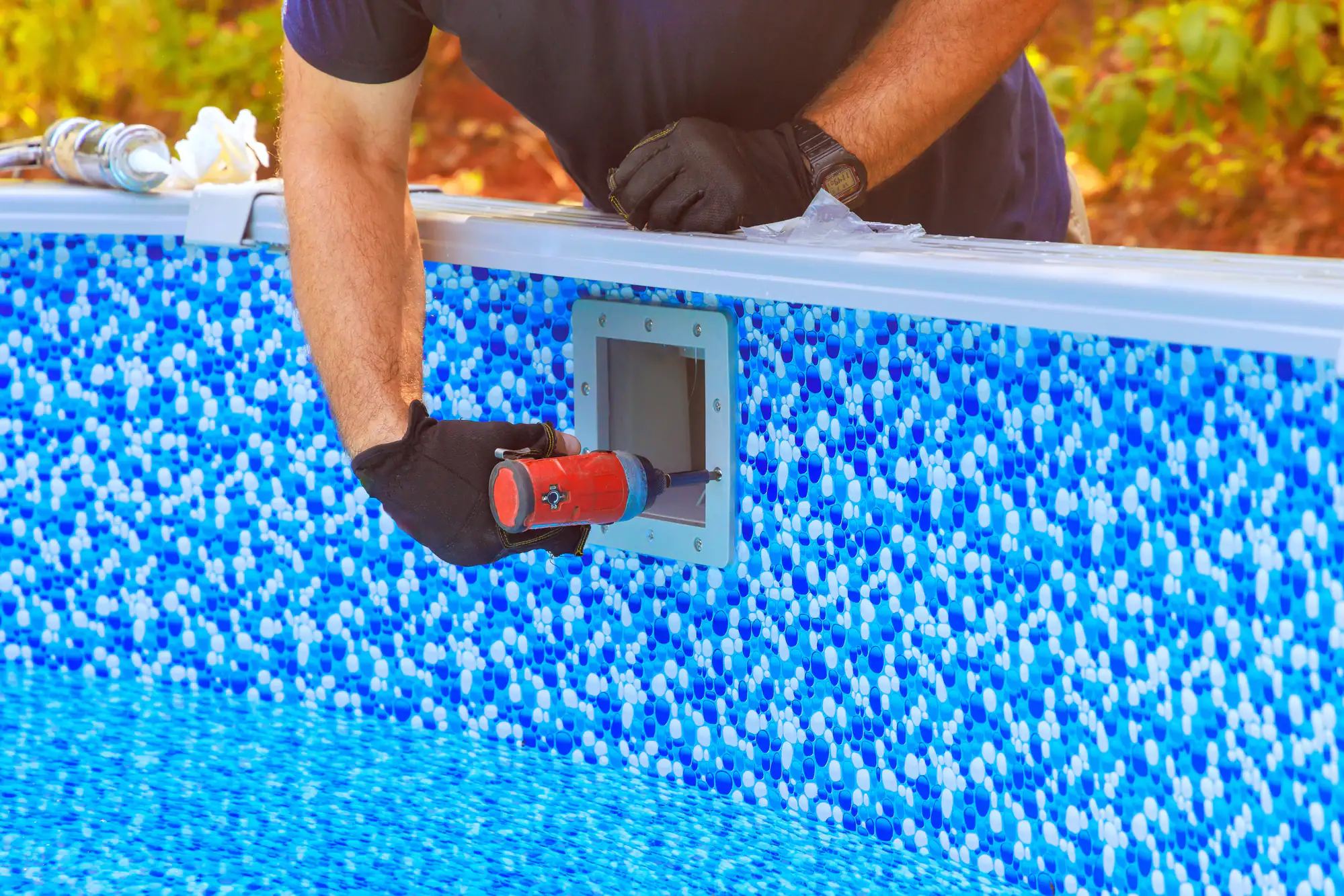 A person wearing black gloves applies sealant with a caulking gun around a pool skimmer opening on the patterned blue wall of an above-ground swimming pool.