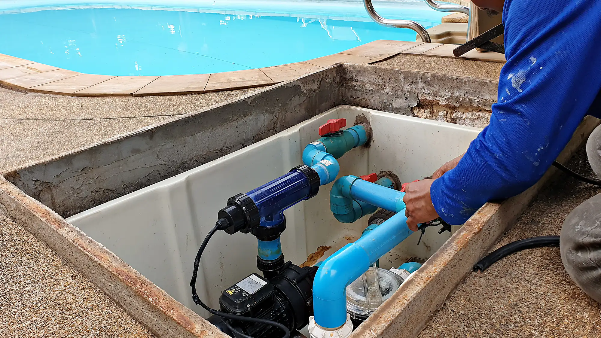 A person repairs blue PVC pipes and pool equipment inside a rectangular maintenance hatch next to a swimming pool.