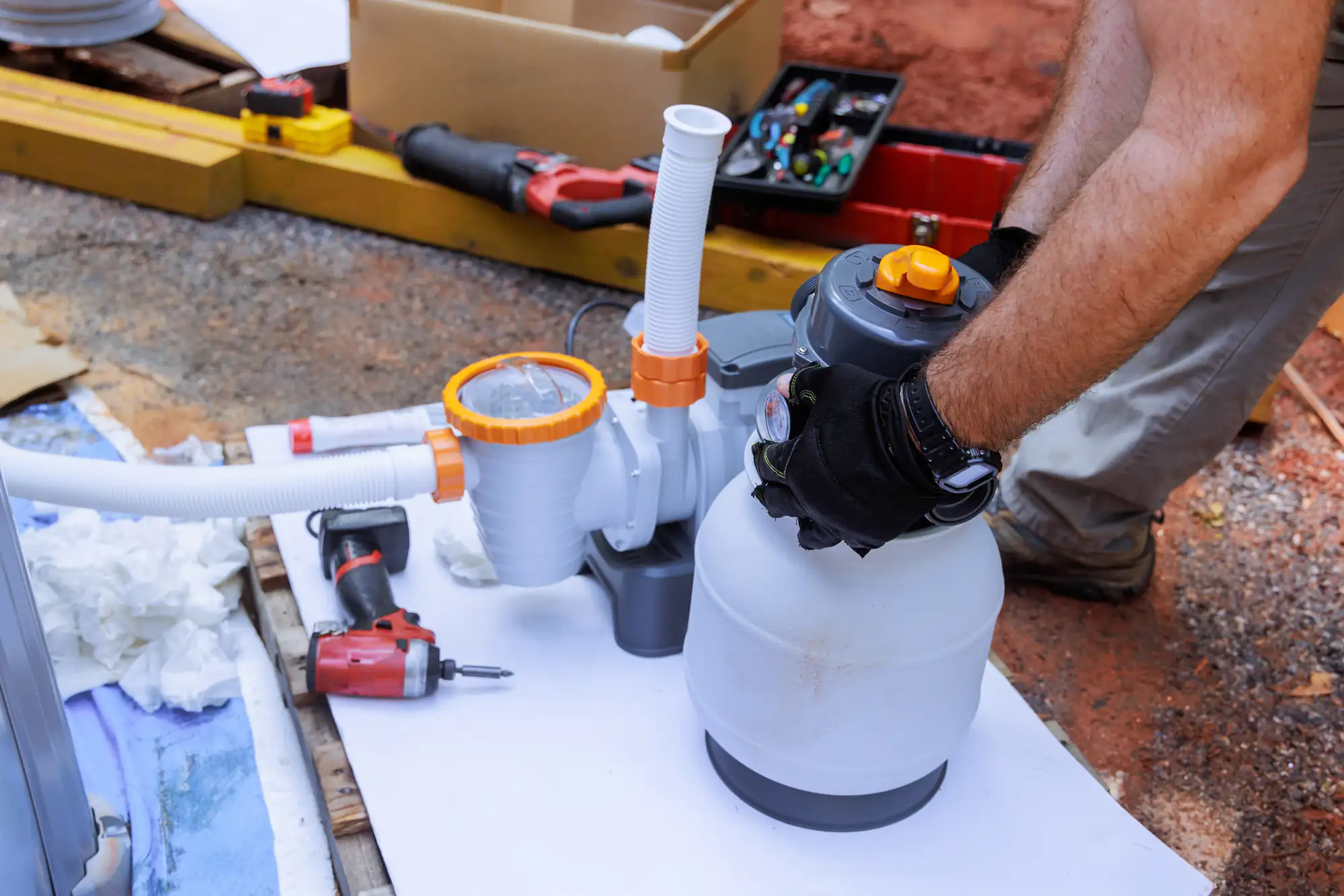 A person wearing gloves assembles or repairs a pool filter pump system outdoors, with tools and parts scattered around on the ground.
