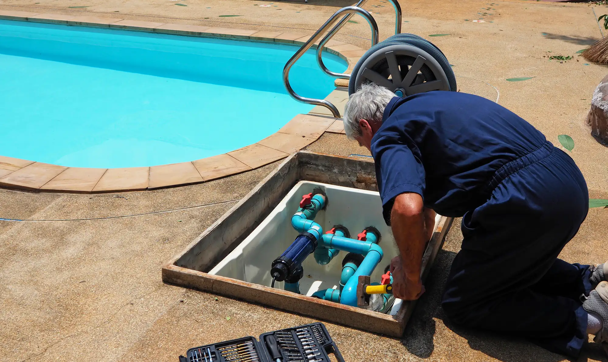 A man in blue coveralls kneels beside a pool, working on pipes and valves in an open equipment hatch. A toolbox and tools are nearby on the pool deck.