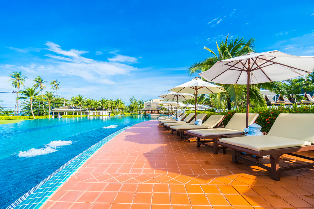 Lounge chairs with white umbrellas line the edge of a large outdoor swimming pool surrounded by palm trees under a bright blue sky.