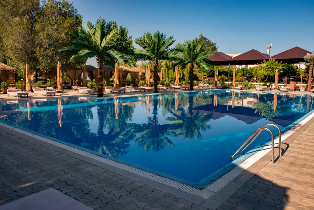 A large outdoor swimming pool with clear blue water reflects palm trees and a bright sky. Lounge chairs and umbrellas line the poolside on a tiled deck, with greenery and shaded pavilions in the background.