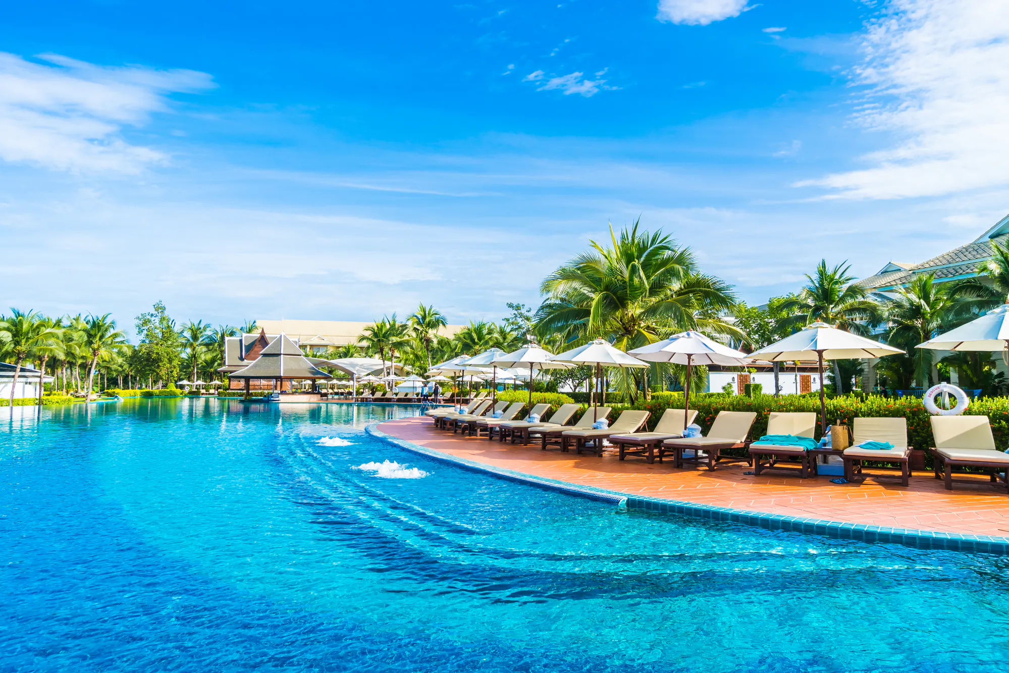 A large outdoor swimming pool with clear blue water, surrounded by lounge chairs and umbrellas, tropical palm trees, and resort buildings under a bright blue sky.
